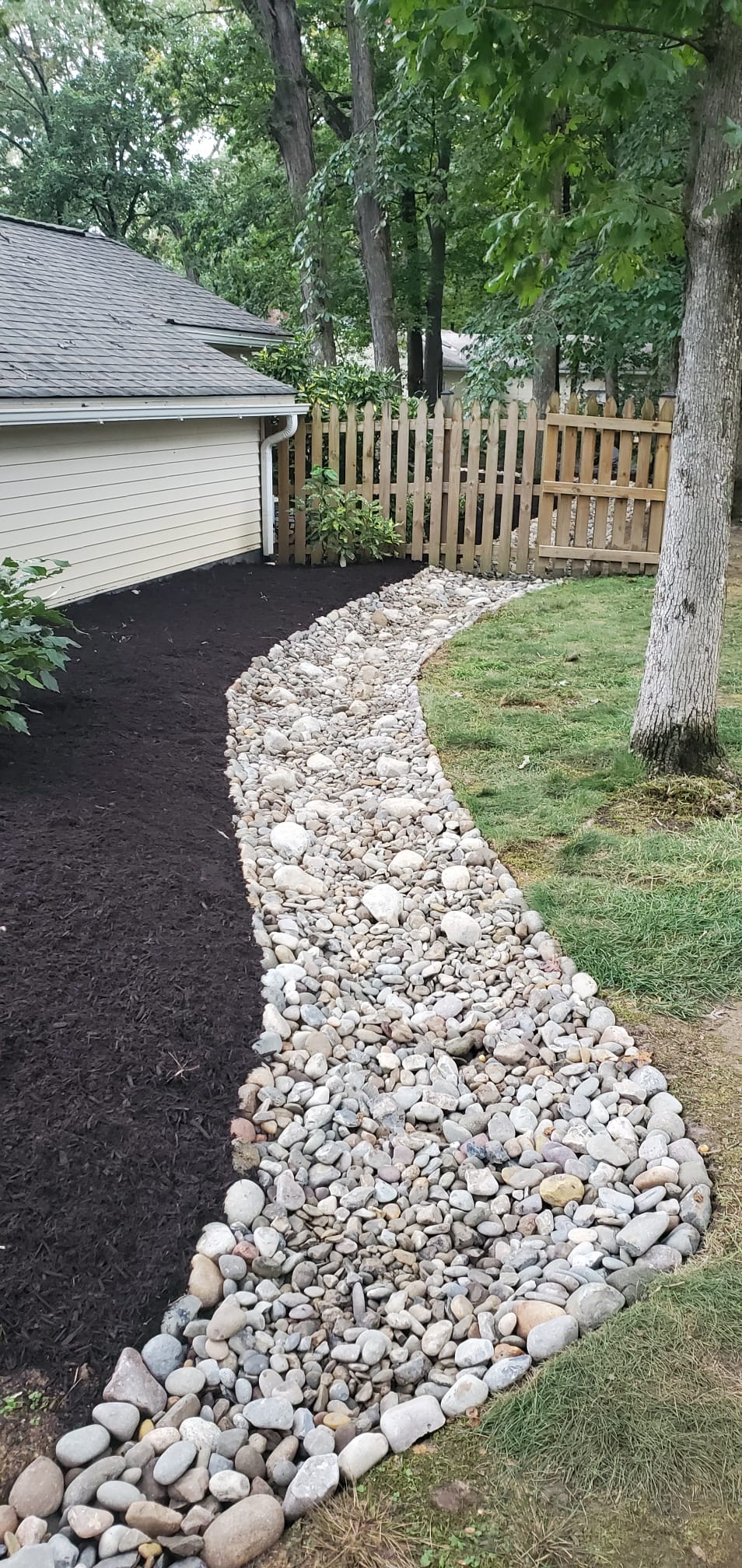Finished stone-lined residential drainage swale beside a garage and planting bed